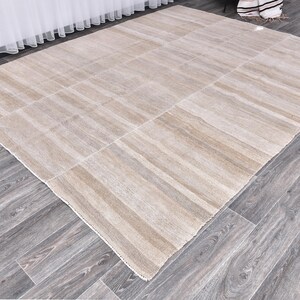 Pu&ograve; includere: Un tappeto beige e bianco a strisce con un leggero bagliore. Il tappeto &egrave; steso su un pavimento in legno.