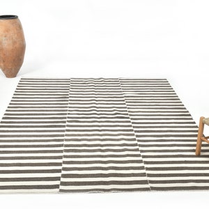 May include: A striped rug with brown and white stripes. The rug is laid out on a white surface with a clay pot and a wooden stool in the background.