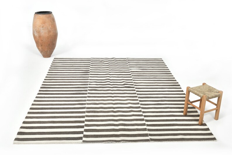 May include: A striped rug with brown and white stripes. The rug is laid out on a white surface with a clay pot and a wooden stool in the background.