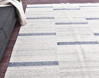 May include: A rectangular, off-white rug with navy blue horizontal stripes. The rug is made of a woven material and is placed on a hardwood floor. The rug is next to a gray sofa with a cream-colored throw blanket.
