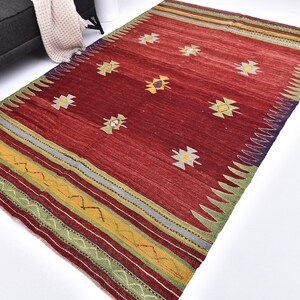 May include: A rectangular area rug featuring a deep red field with scattered geometric motifs in yellow, white, and blue. The borders are decorated with horizontal stripes in green, yellow, and red, accented by a zigzag pattern along the edges.