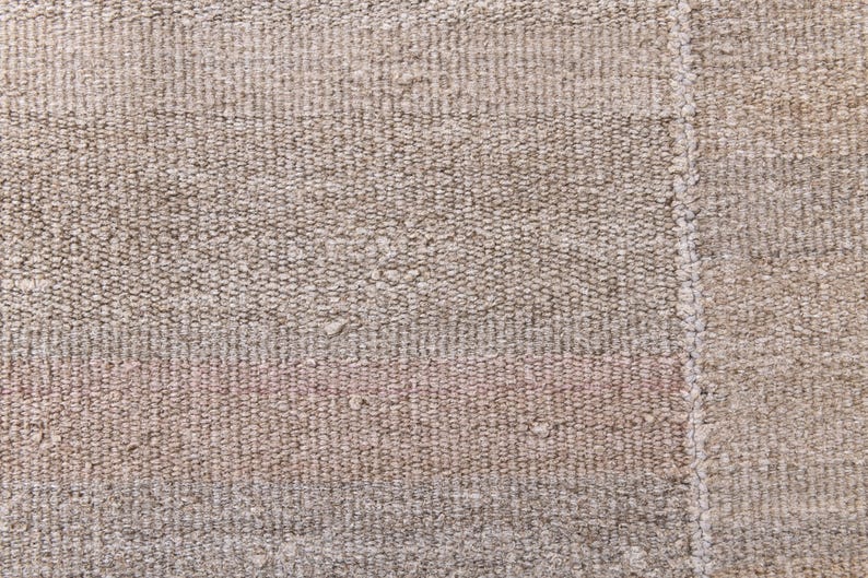 Pu&ograve; includere: Primo piano di un tappeto beige tessuto con una texture sottile. Il tappeto presenta una striscia leggermente pi&ugrave; scura che attraversa orizzontalmente il centro.