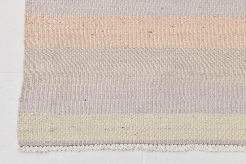 K&ouml;nnte beinhalten: Ein gewebter Teppich mit einem gestreiften Muster in Hellrosa, Creme und Hellgrau. Der Teppich hat einen Fransenrand.