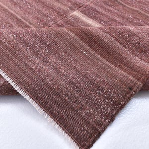 May include: A close-up of a folded, woven rug in shades of brown and mauve. The rug has a textured surface with visible fibers and a white fringe along the edge. The rug is folded to reveal the underside.