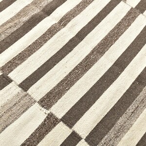 Op de afbeelding: Close-up van een geweven tapijt met een streepjespatroon. Het tapijt heeft afwisselende banden van lichtbeige en donkerbruin. Het ontwerp bestaat uit rechthoekige patches, waardoor een textuur ontstaat.