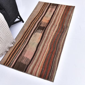 May include: A rectangular rug with a striped pattern in shades of brown, beige, and orange. The rug has a woven texture and is placed on a white surface. The rug's design features vertical stripes of varying widths and colors.