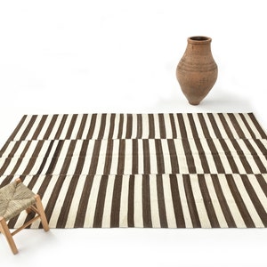 May include: A brown and white striped rug with a woven stool in the foreground and a terracotta vase in the background.