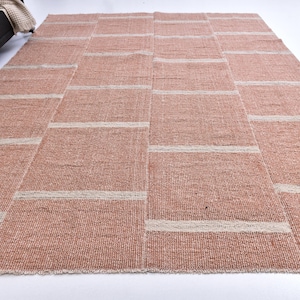 Op de afbeelding: Een groot, rechthoekig vloerkleed met een patchwork ontwerp. Het vloerkleed heeft een warme, roodbruine kleur met horizontale banden in een lichtere, neutrale tint. De textuur lijkt geweven, wat wijst op een natuurlijk vezelmateriaal.