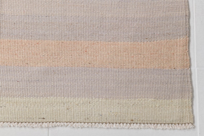 K&ouml;nnte beinhalten: Ein gewebter Teppich mit einem Streifenmuster in Beige-, Hellrosa- und Graut&ouml;nen. Der Teppich hat einen Fransenrand.