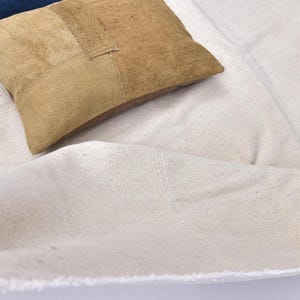 Könnte beinhalten: Nahaufnahme eines cremefarbenen Stoffes mit ausgefranstem Rand und zwei Kissen. Ein Kissen ist beige und geflickt, das andere ist blau. Der Stoff ist drapiert und strukturiert.