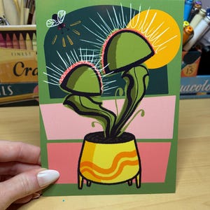 May include: A botanical-themed greeting card featuring a Venus flytrap illustration. The card showcases a green flytrap with red accents, a yellow pot, and a fly. The background includes green, pink, and yellow sections.