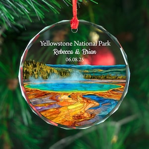 May include: A clear, round Christmas ornament with a colorful Yellowstone National Park design. The ornament features a landscape with a blue lake, yellow and orange thermal features, and the text "Yellowstone National Park Rebecca & Brian 06.08.25". A red ribbon is attached.