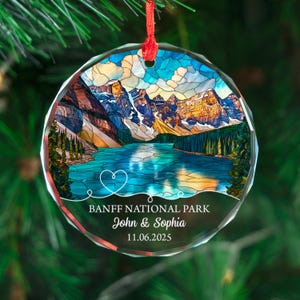 May include: A round, clear glass Christmas ornament with a scenic view of Banff National Park. The image features a lake, mountains, and trees, with the text "BANFF NATIONAL PARK" and names "John & Sophia" with a date.