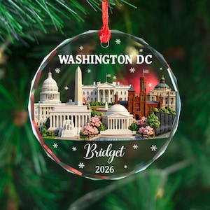 May include: A clear, round Christmas ornament with a Washington DC cityscape design. The ornament features the text "WASHINGTON DC", "Bridget", and "2026". The ornament is hanging from a red ribbon against a green background.