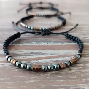 May include: A set of three adjustable bracelets featuring black braided cord, metallic copper-colored cylindrical beads, and polished dark gray spherical beads arranged in a repeating pattern along the front of each piece.
