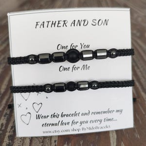 May include: Two black cord bracelets with silver and black beads are displayed on a white card. The card reads "FATHER AND SON" and "One for You, One for Me." The text "Wear this bracelet and remember my eternal love for you every time..." is also visible.