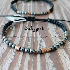 May include: Two black braided bracelets with metallic beads and copper-colored accents. The top bracelet has the word "Daddy" above it, and the bottom bracelet has the word "Babygirl" above it.