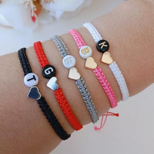 May include: Five braided cord bracelets in black, red, gray, pink, and white. Each bracelet features a single round bead with a letter—T, G, B, B, or K—and a small metallic heart charm. The letters are printed in contrasting colors on the beads.