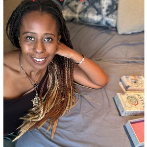 May include: A woman with long braided hair wearing a black top and a silver necklace sits on a bed with a deck of Astro Oracle cards and a pink box.