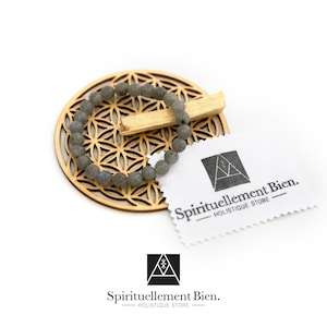 Może przedstawiać: Bransoletka z szarego labradoritu na drewnianym mandali Kwiatu Życia z patyczkiem palo santo. Bransoletka leży na białym tle z białą etykietą, na której widnieje napis "Spirituellement Bien. -HOLISTIQUE STORE- "