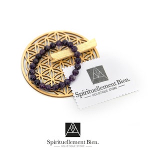 May include: A purple amethyst gemstone bracelet on a wooden mandala-shaped stand. The bracelet is made of small, round beads. The stand has a flower of life design. The image also includes a white card with the text "Spirituellement Bien. - HOLISTIQUE STORE -"