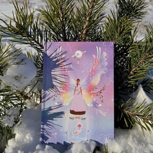 Könnte beinhalten: Ein Hardcover-Journal mit einer farbenfrohen Illustration einer Frau mit Engelsflügeln, vor einer schneebedeckten Landschaft und Kiefernzweigen. Das Cover-Artwork zeigt Pastellfarben und ein skurriles Design.
