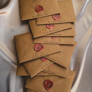May include: A stack of brown kraft paper envelopes, each featuring small gold star illustrations and the handwritten text "DO YOU BELIEVE IN MAGIC?". Each envelope is sealed with a circular red wax seal embossed with a circular logo design.