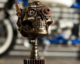 Può includere: Una statuetta di teschio in stile steampunk con ingranaggi e ruote dentate, una finitura in bronzo e un'unica lente oculare. Il teschio è montato su un supporto decorativo con una base a forma di ingranaggio.