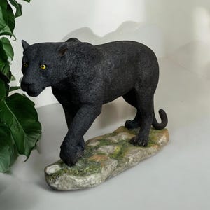 Può includere: Figurina di pantera nera in piedi su una base rocciosa con muschio verde. La pantera è in posizione di camminata con la testa girata di lato.