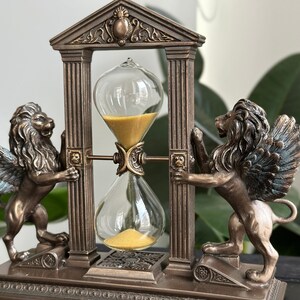 May include: A bronze hourglass with winged lion statues on either side, set within a decorative archway. The hourglass is filled with golden sand and has a clear glass bulb.
