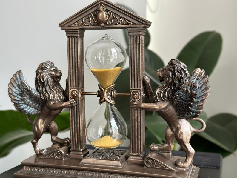 May include: A bronze hourglass with winged lion statues on either side, set within a decorative archway. The hourglass is filled with golden sand and has a clear glass bulb.