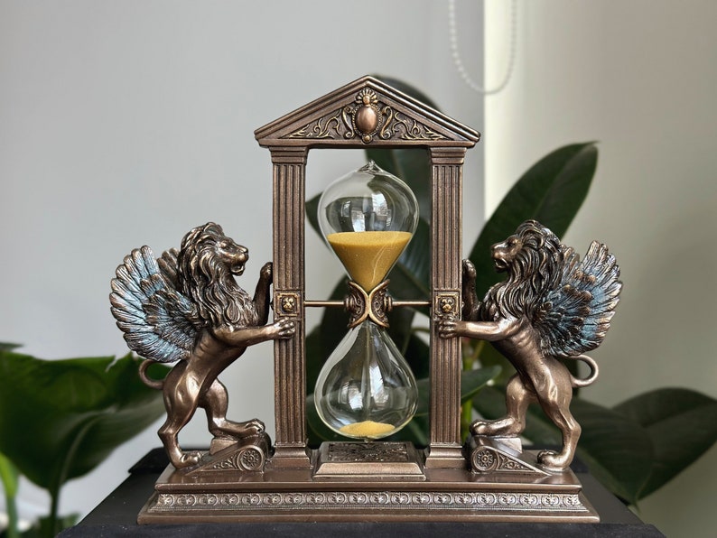 May include: A bronze hourglass with winged lion sculptures on each side, set within a decorative archway. The hourglass is filled with golden sand and sits on a base with intricate details.