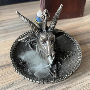 May include: A silver metal incense burner shaped like a goat's head with horns and a pentagram on its forehead. Smoke is rising from the burner, which is sitting on a circular base with a decorative border.