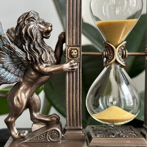 May include: A winged lion statue holding up a decorative hourglass with gold sand. The hourglass is framed by two pillars with ornate details. The base is made of dark brown wood.
