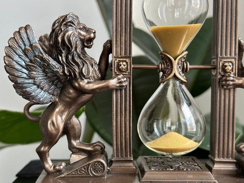 May include: A winged lion statue holding up a decorative hourglass with gold sand. The hourglass is framed by two pillars with ornate details. The base is made of dark brown wood.