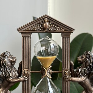May include: A bronze hourglass with winged lion statues on either side. The hourglass is framed by a decorative archway with ornate details. The sand is golden and the glass is clear.