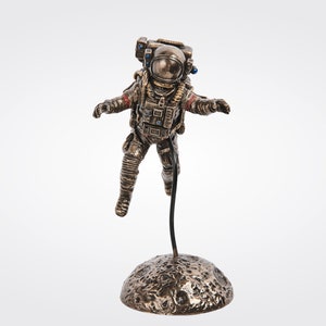 May include: A bronze figurine of an astronaut in a space suit, floating in mid-air above a small, cratered moon-like base.