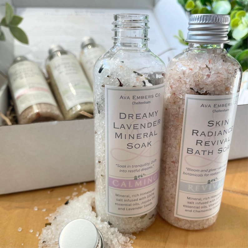 May include: Two clear glass bottles filled with bath salts, labeled "Dreamy Lavender Mineral Soak" and "Skin Radiance Revival Bath Soa". The bottles are in a gift box with other bath products. The labels have text and a soft color palette.