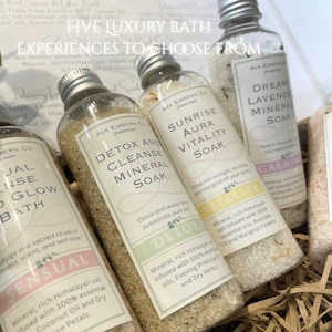 May include: A collection of bath soaks in clear bottles with silver caps. The labels read "Detox and Cleanse Mineral Soak", "Sunrise Aura Vitality Soak", and "Dreamy Lavender Mineral Soak". The text "Five Luxury Bath Experiences to Choose From" is at the top.