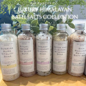 May include: A collection of five clear glass bottles filled with bath salts. Each bottle has a silver cap and a label with text. The labels read "Sunrise Aura Vitality Soak", "Ritual Sense and Glow Bath", "Dreamy Lavender Mineral Soak", "Detox and Cleanse Mineral Soak", and "Skin Radiance Revival Bath Soak".