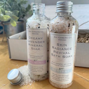 May include: Two clear glass bottles filled with mineral bath soak. One is labeled "Dreamy Lavender Mineral Soak" and the other "Skin Radiance Revival Bath Soak." Both have silver caps and white labels with black text. The bottles are on a wooden surface.