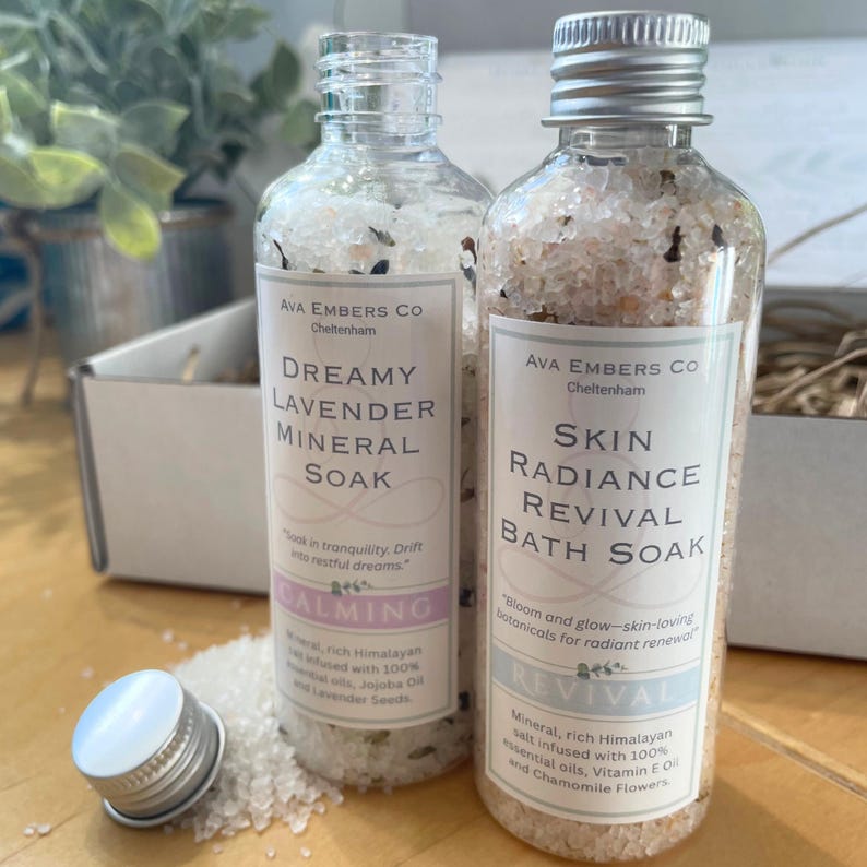 May include: Two clear glass bottles filled with mineral bath soak. One is labeled "Dreamy Lavender Mineral Soak" and the other "Skin Radiance Revival Bath Soak." Both have silver caps and white labels with black text. The bottles are on a wooden surface.