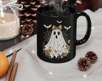 May include: Black ceramic mug featuring a Halloween ghost design. The ghost is draped in a white lace sheet with a gold bow, surrounded by bats, spiderwebs, and flowers. The mug has a curved handle and is filled with a dark liquid.