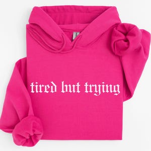 May include: A bright pink hoodie with a white graphic that says "tired but trying" in a gothic font.