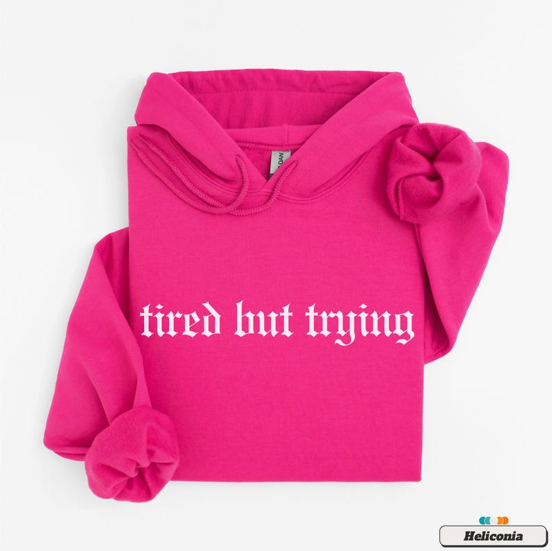 May include: A bright pink hoodie with a white graphic that says "tired but trying" in a gothic font.