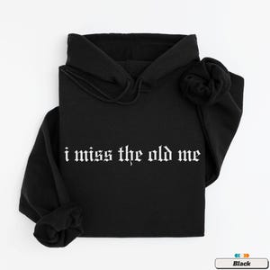 Puede incluir: Sudadera con capucha negra con el texto "i miss the old me" en fuente g&oacute;tica blanca.