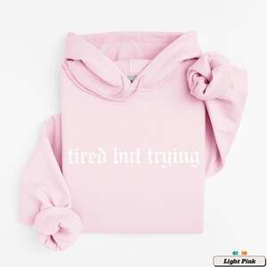 May include: A light pink hoodie with the text "tired but trying" in white gothic font.