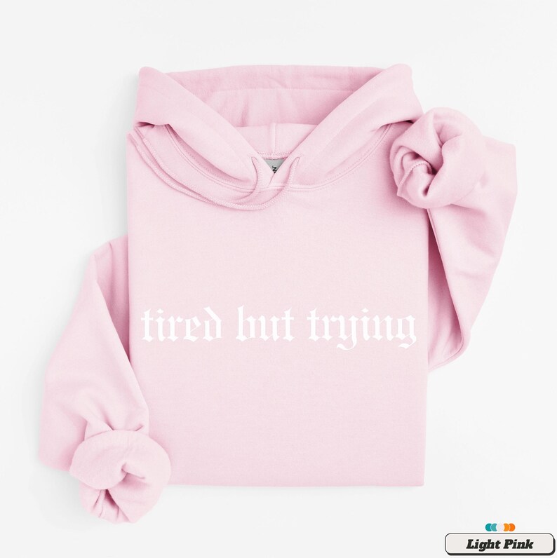 May include: A light pink hoodie with the text "tired but trying" in white gothic font.