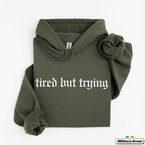 May include: A green hooded sweatshirt with the text "tired but trying" in white gothic font.