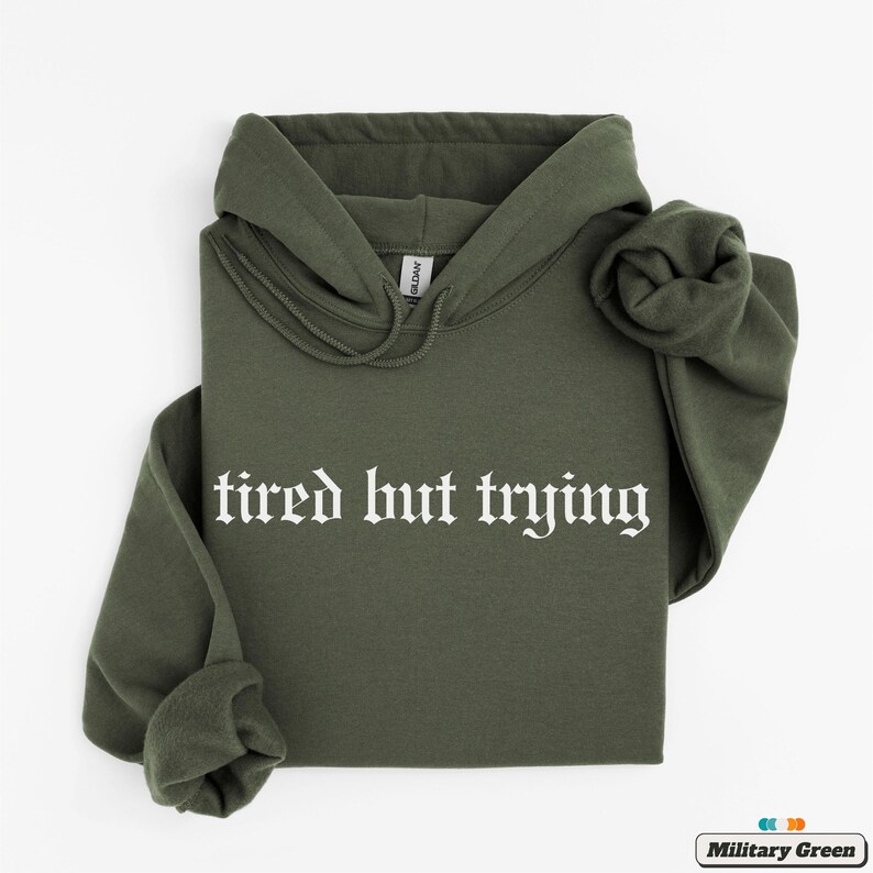 May include: A green hooded sweatshirt with the text "tired but trying" in white gothic font.